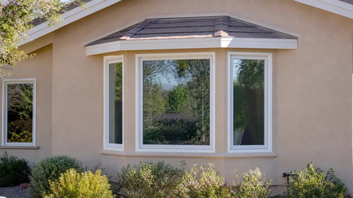 A tan stucco house wall with a three-panel bay window under a small hip roof, shrubs planted below.